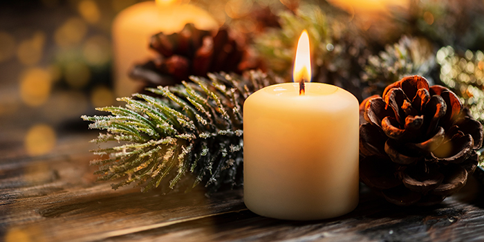 Single column white candle with soft flame in front of holiday greenery and pinecone